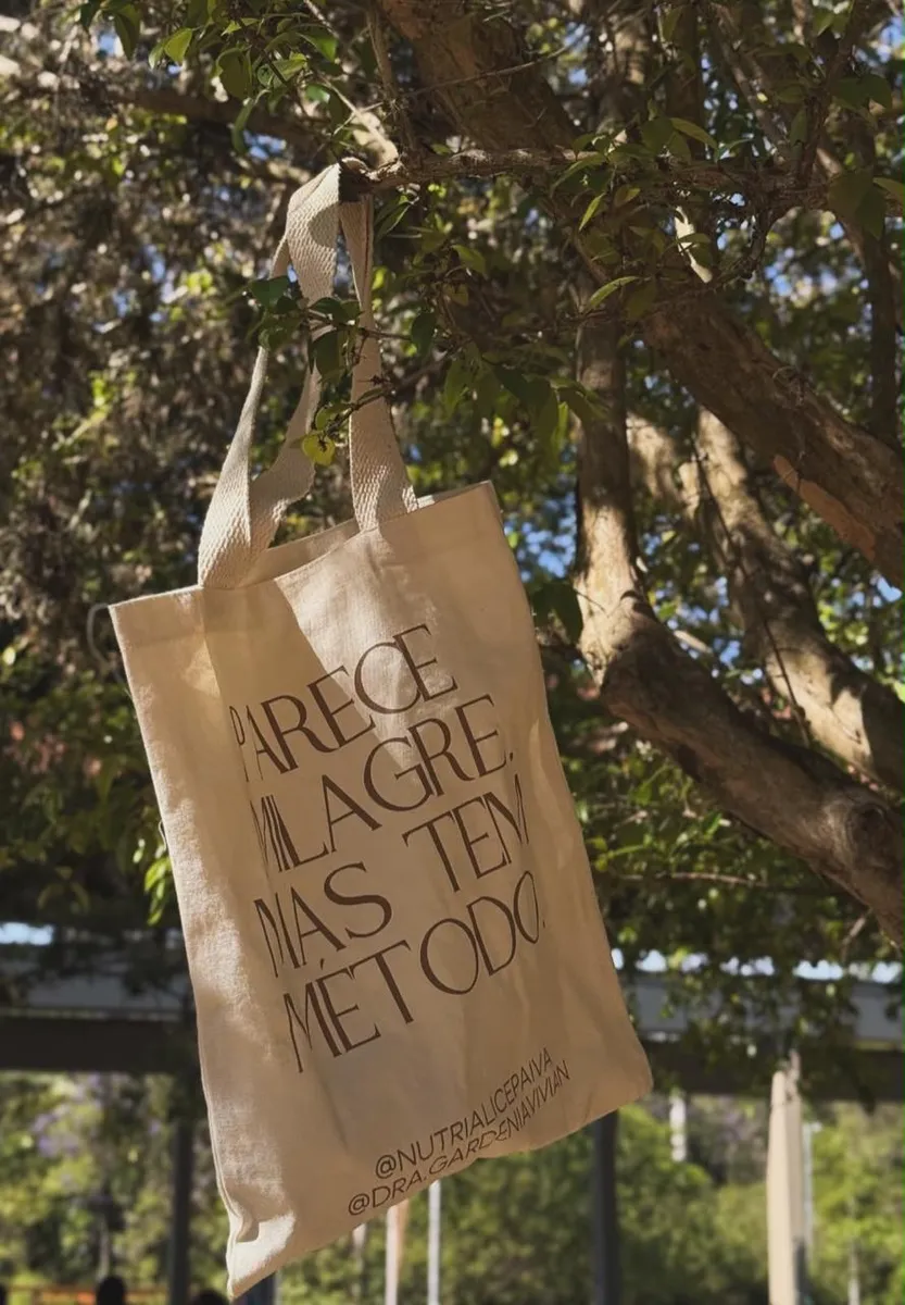 Ecobag do evento com o lettering Parece Milagre Mas Tem Método pendurada em uma árvore